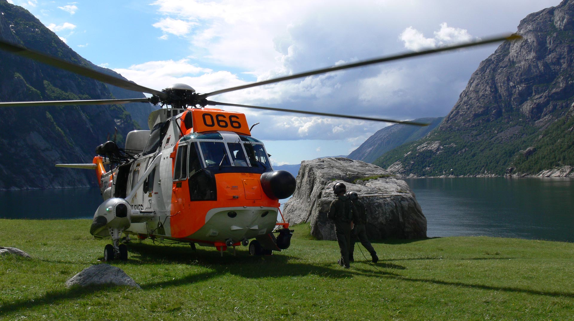 Seaking helikopter og to personer med fjell og fjord i bakgrunn.
