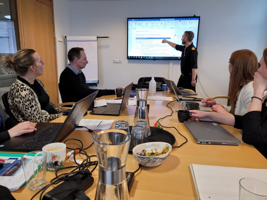 Bilde av mennesker ved et møtebord mens en person presenterer noe ved en skjerm