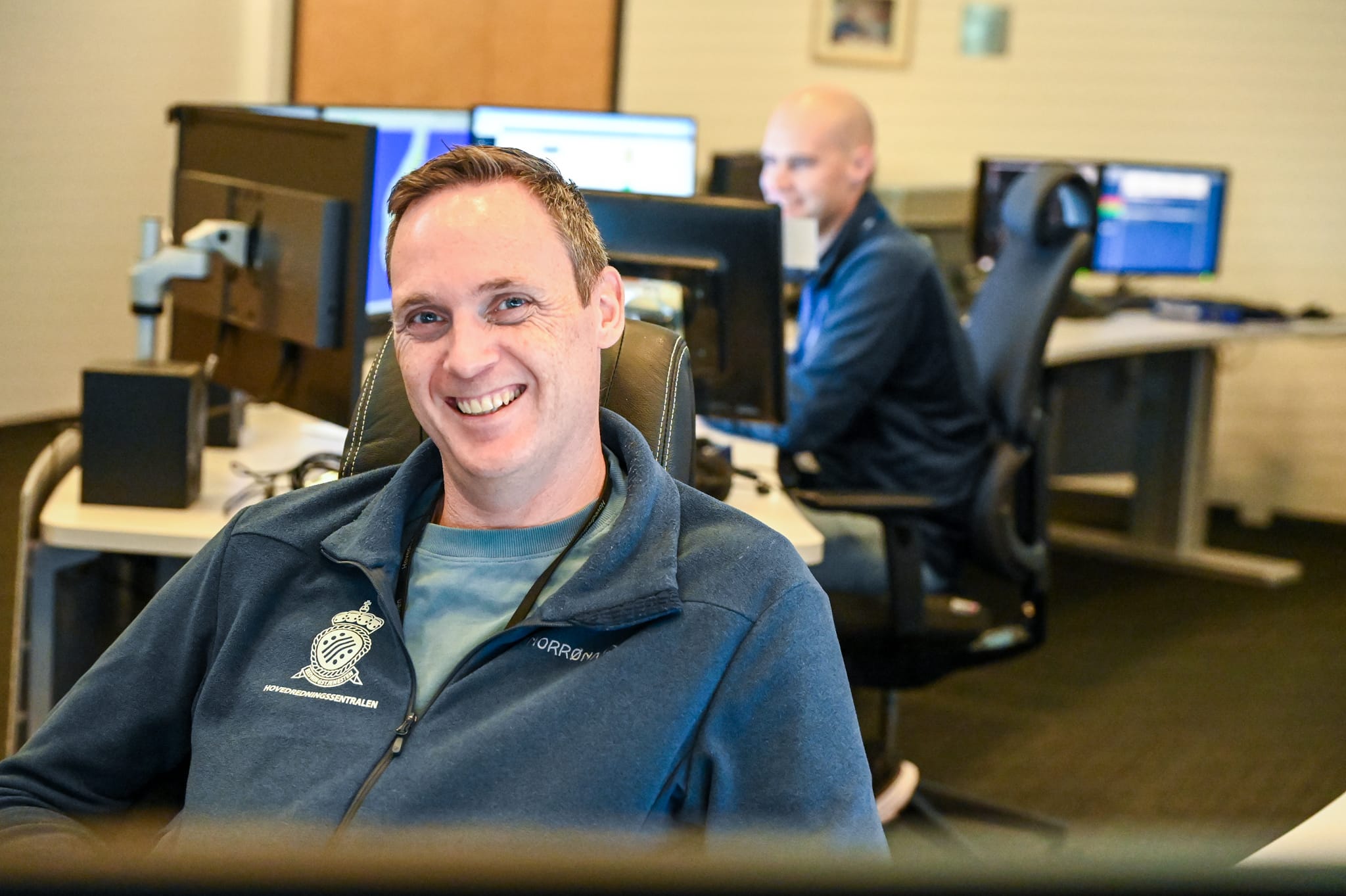 Mann smiler, sitter innendørs, annen mann er uskarp i bakgrunnen