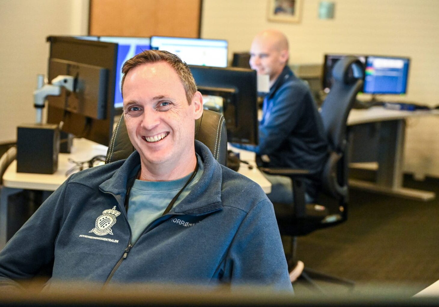 Jan Eirk Skorpe Mann smiler, sitter innendørs, annen mann er uskarp i bakgrunnen