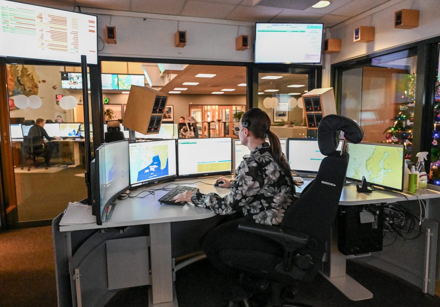 Trine T-J Kvinne sitter ved pult med mange dataskjermer.